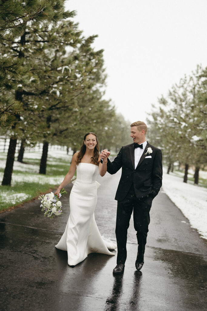 Bride and groom smiling at each other while walking down a snowy tree-lined road