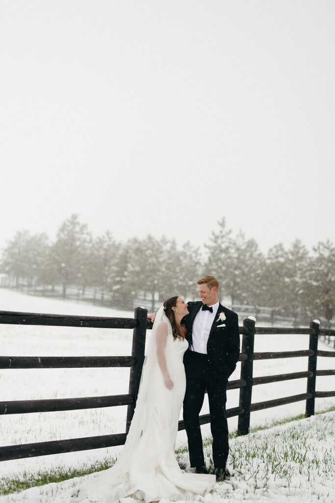 Bride and groom leaning against wooden fence surrounded by fresh snowfall