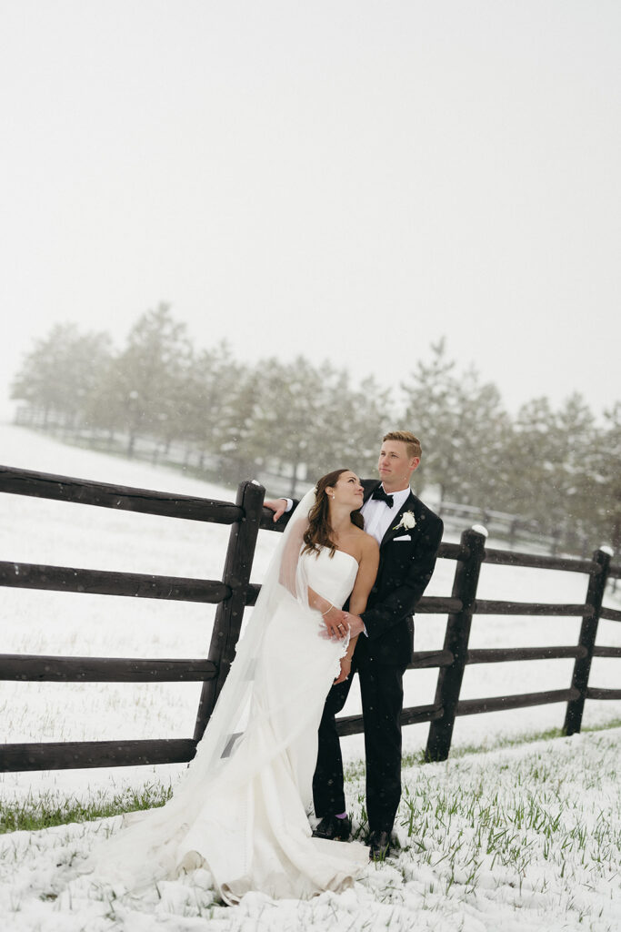 Bride and groom leaning against wooden fence surrounded by fresh snowfall