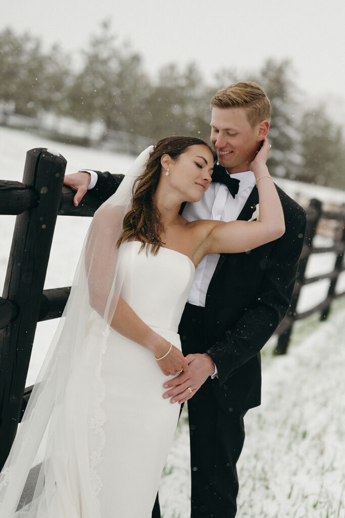 Bride and groom leaning against wooden fence surrounded by fresh snowfall