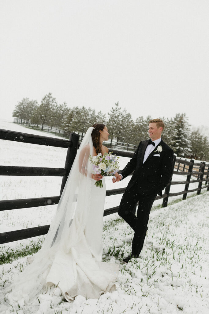 Bride and groom leaning against wooden fence surrounded by fresh snowfall